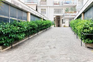 A narrow stone-paved corridor flanked by green hedges and a multi-story building with balconies. - Appartamento luminoso a San Pietro (Roma)