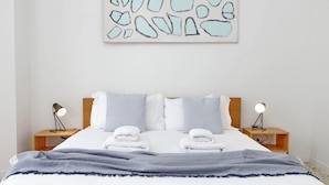 Una cama bien hecha con sábanas blancas y dos almohadas, flanqueada por dos mesitas de noche con lámparas.