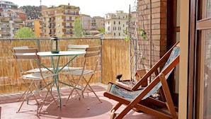 Un balcón con una mesa y sillas, una tumbona y vistas de edificios circundantes.