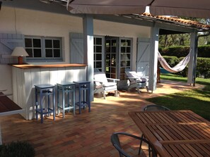 Une terrasse couverte avec une table et des chaises en bois, un hamac et un coin bar avec des tabourets.