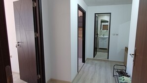 A narrow hallway with a wooden door on the left, a metal rack with shoes on the right, and a bathroom visible through an open door in the background. - Perfect 1-bedroom apartment in adorable Pamporovo for your getaway (Pamporovo)