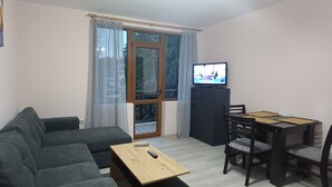 A compact living space with a sofa, a small dining table with chairs, a television, and a sliding glass door leading to a balcony. - Perfect 1-bedroom apartment in adorable Pamporovo for your getaway (Pamporovo)