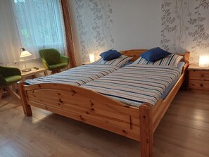 Een houten bed met gestreepte beddengoed, een groene stoel, een nachtkastje met een lamp, en behang met bloemen.