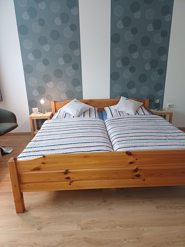 A wooden bed with striped bedding, two bedside tables, a lamp, and a chair in a room with patterned wallpaper. - Room “Double Room 3” with Shared Terrace, Garden, and Wi-Fi (Kirchhundem)
