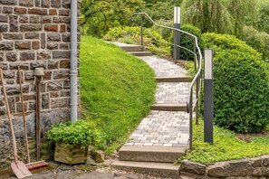 Eine Steinmauer, ein Pflasterweg aus Ziegeln, Metallgeländer und grüne Hecken.