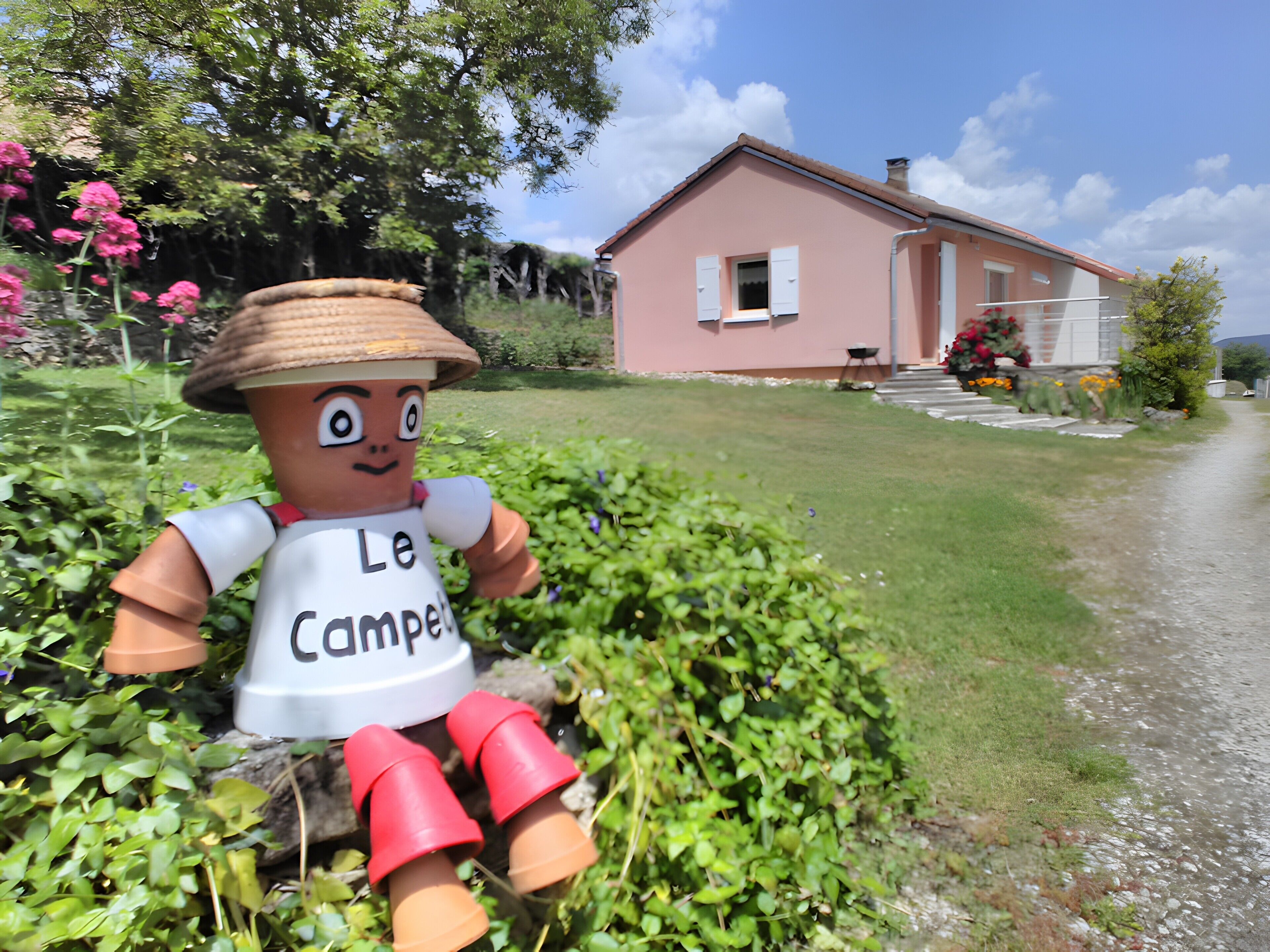 Une maison à l’extérieur rose, un jardin verdoyant et fleuri, et une statue représentant une personne portant un chapeau et un t-shirt sur lequel on peut lire « Le Campe ».