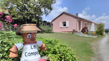Une maison à l’extérieur rose, un jardin verdoyant et fleuri, et une statue représentant une personne portant un chapeau et un t-shirt sur lequel on peut lire « Le Campe ».