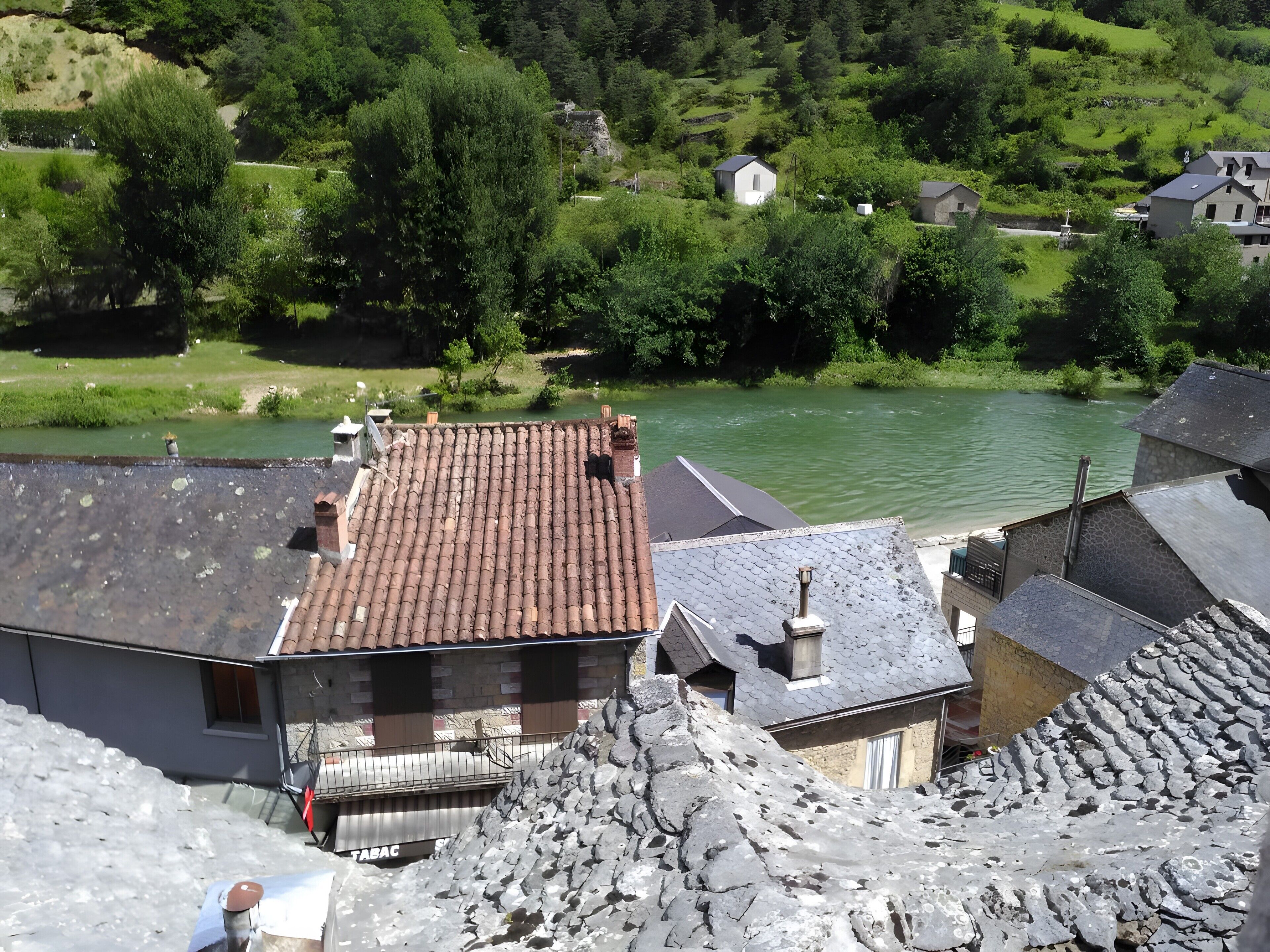 Un bâtiment traditionnel en pierre, avec un toit en tuiles, situé au bord d’une rivière, entouré de verdure et d’autres bâtiments.