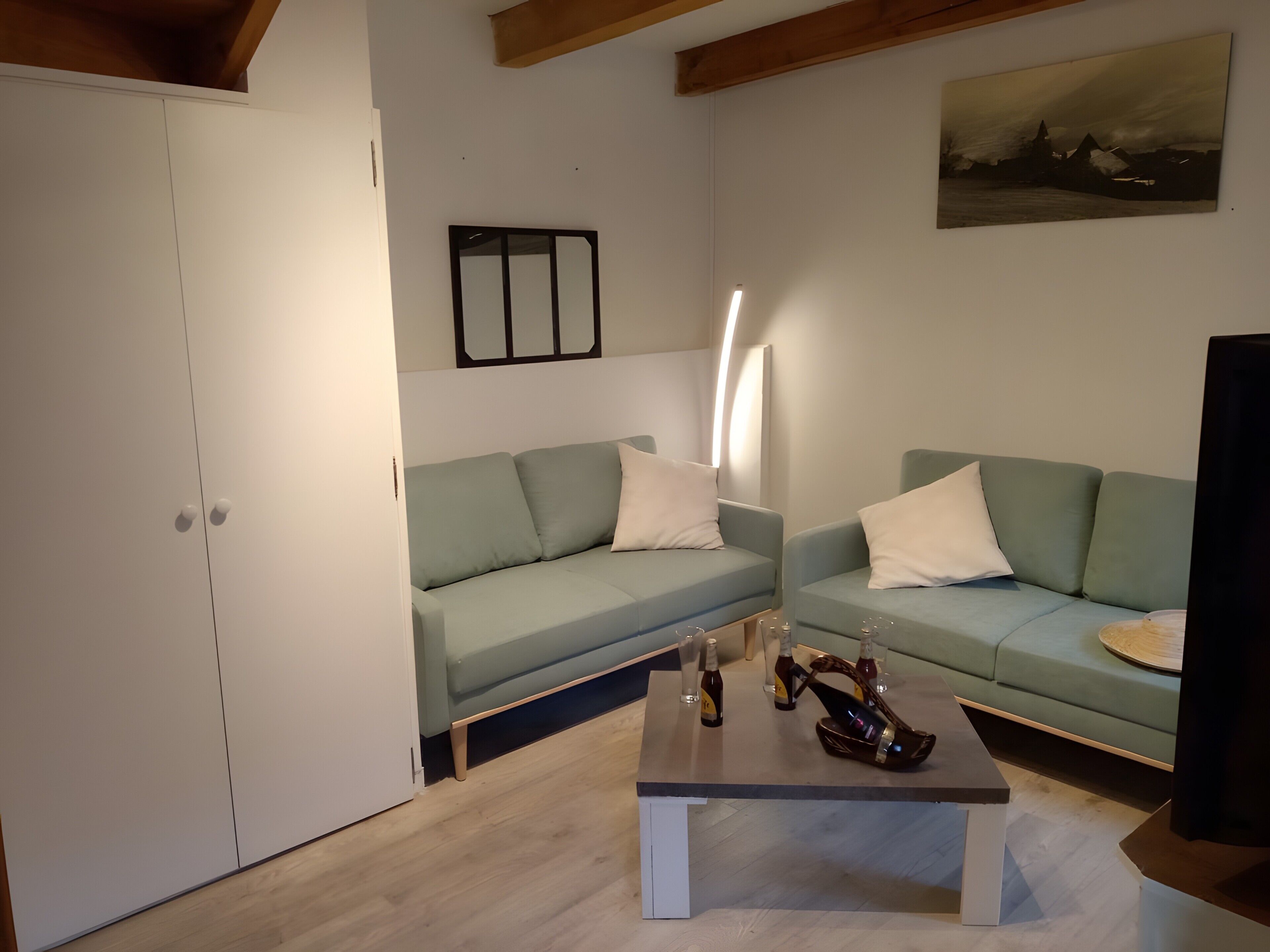 Un salon avec deux canapés vert clair, une table basse sur laquelle sont posées des bouteilles, un tableau au mur et un plafond en bois.