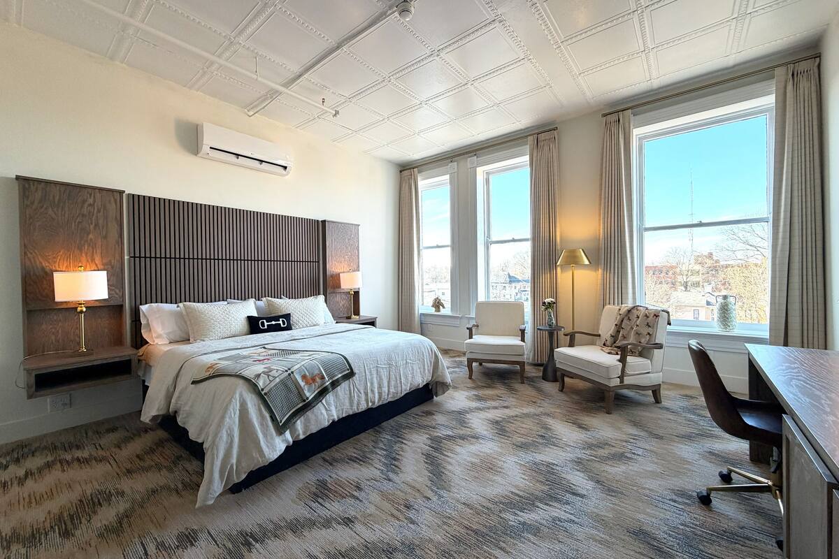 A modern hotel room with a large bed, two armchairs, a desk, and a view of the city through large windows.