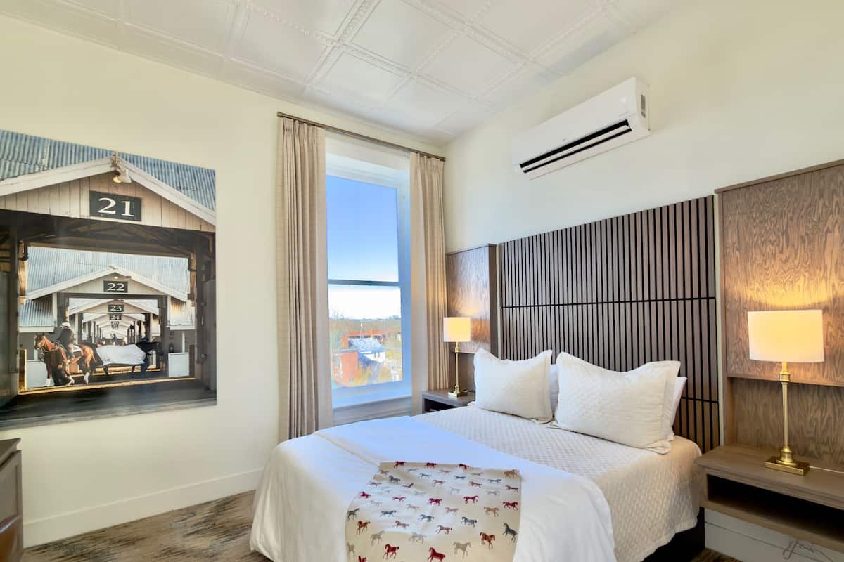 A hotel room with a large bed, a nightstand with a lamp, a window with curtains, and a wall-mounted air conditioner.