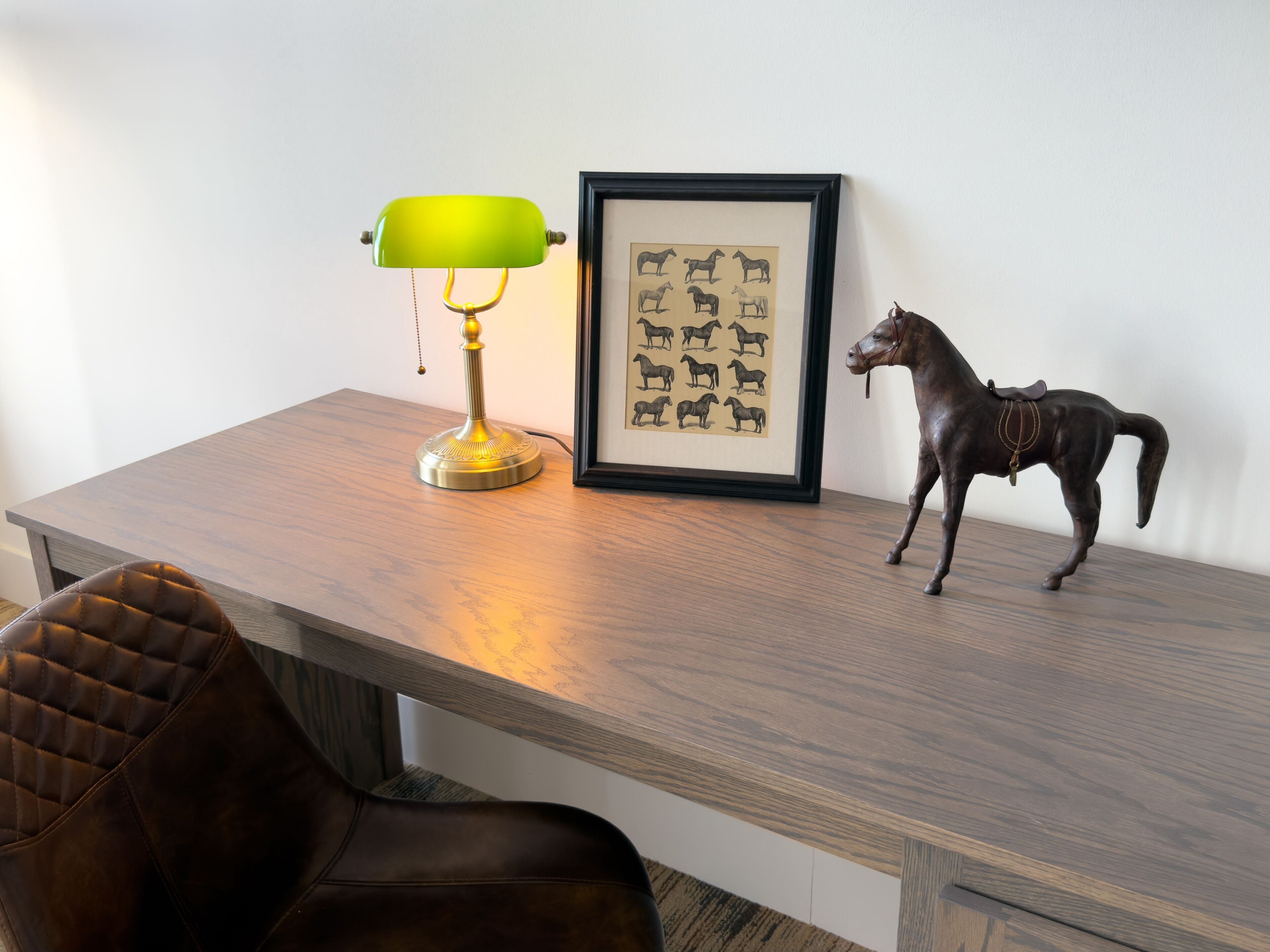 A wooden desk with a green desk lamp, a framed picture of horses, and a horse figurine.