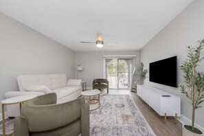 Un salon moderne avec un canapé blanc, un fauteuil vert, un tapis à motifs, un ventilateur de plafond et un téléviseur fixé au mur.