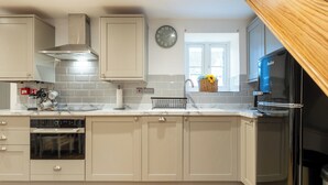 A modern kitchen with beige cabinetry, a built-in oven, a stainless steel range hood, a wall clock, and a window with a basket of flowers. - The Innkeepers Lodge in Cerney Wick (Cirencester)