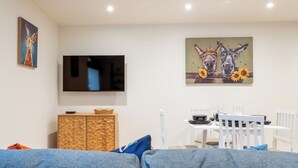 A living room with a TV, a painting of donkeys, a dining table set, and a blue couch. - The Innkeepers Lodge in Cerney Wick (Cirencester)