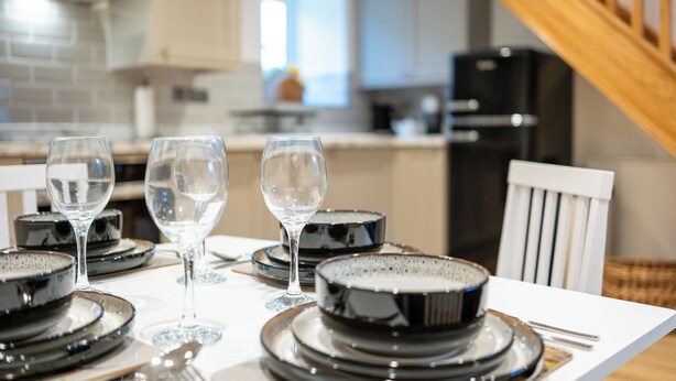 A dining table set with black plates and clear glasses, a white chair, and a kitchen in the background. - The Innkeepers Lodge in Cerney Wick (Cirencester)