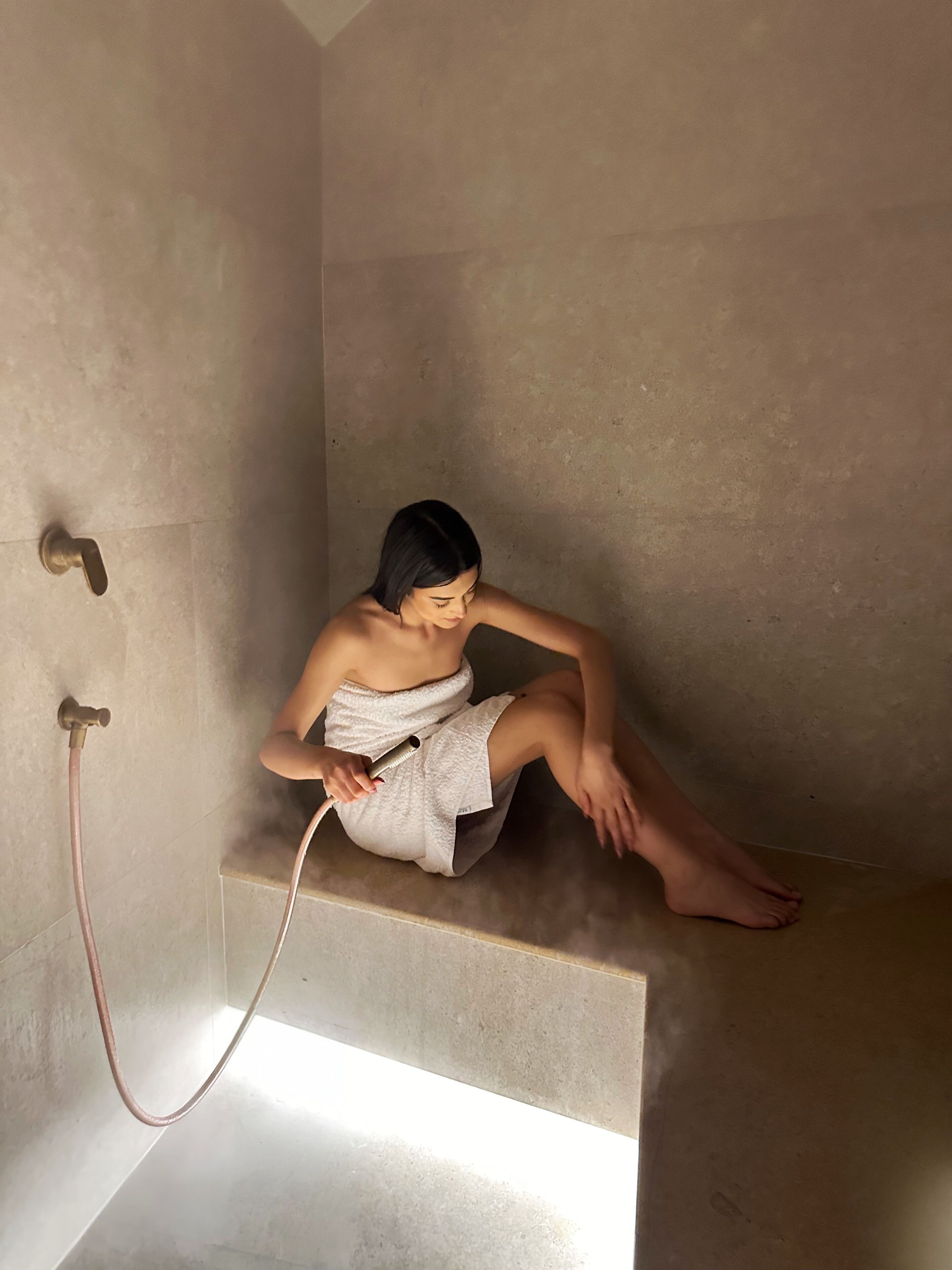 Une personne assise dans un espace de douche, tenant un pommeau de douche à main, une serviette enroulée autour de son corps, et un tuyau d'eau visible.