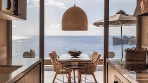 A modern kitchen with a view of the sea, featuring wooden cabinets, a dining area with wicker chairs, and a hanging light fixture.