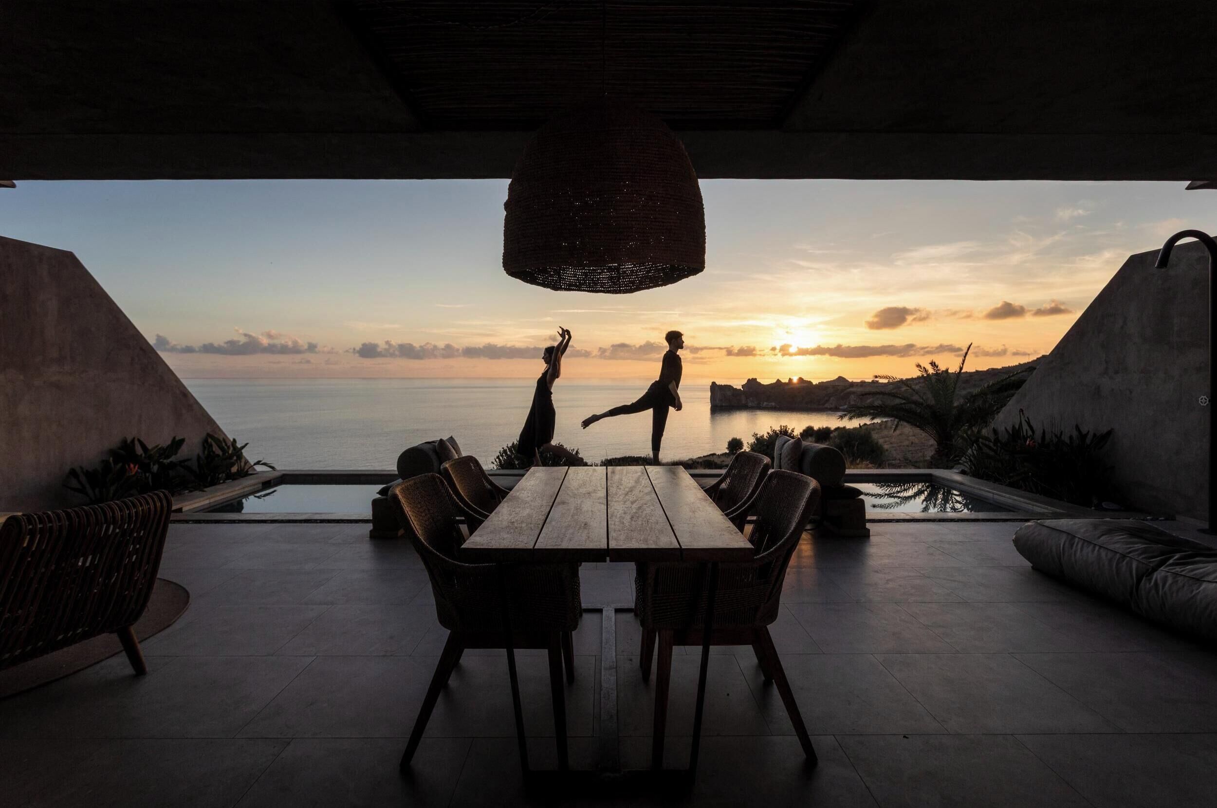 Un área de comedor al aire libre con vistas a la puesta de sol sobre el agua, rodeada de mobiliario moderno y una lámpara colgante.