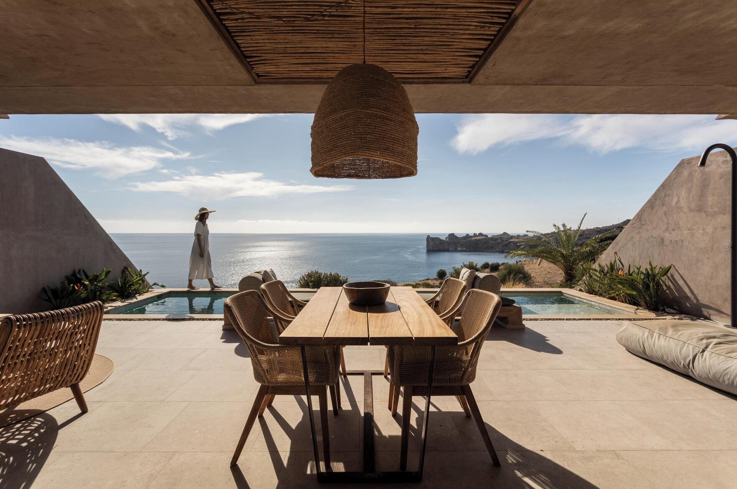 Een zwembadterras met een houten tafel en rieten stoelen, met uitzicht op een schilderachtig zeegezicht.