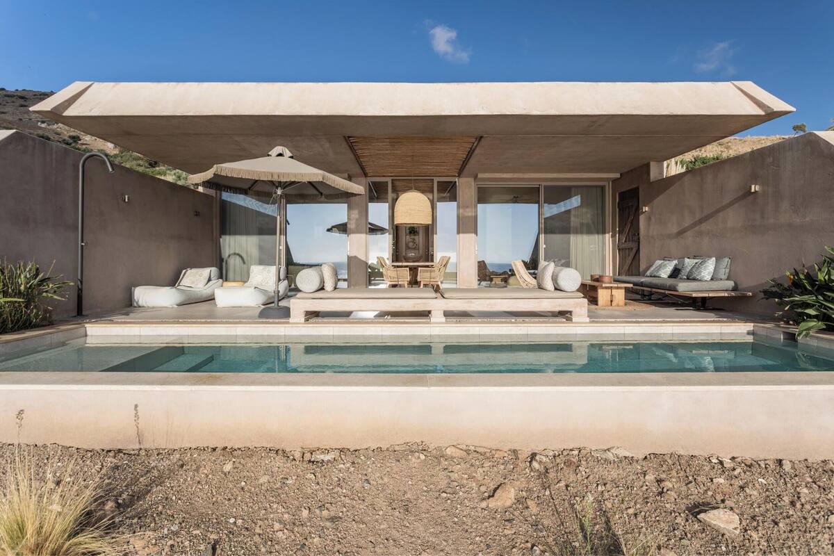 A modern outdoor pool area with a covered seating area, lounge chairs, and a table with umbrellas.