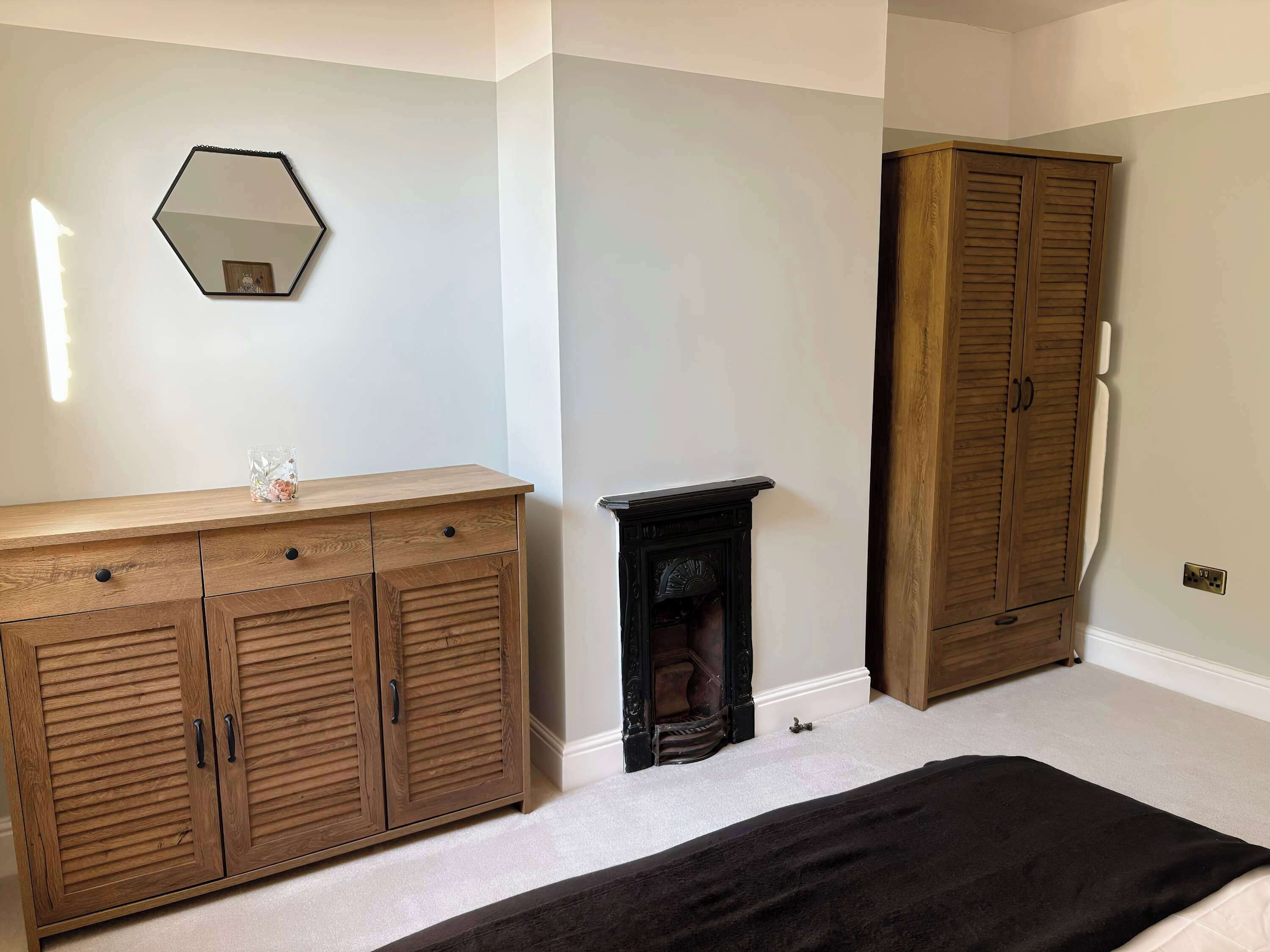 A bedroom with a wooden wardrobe, a dresser, a mirror, and a fireplace.