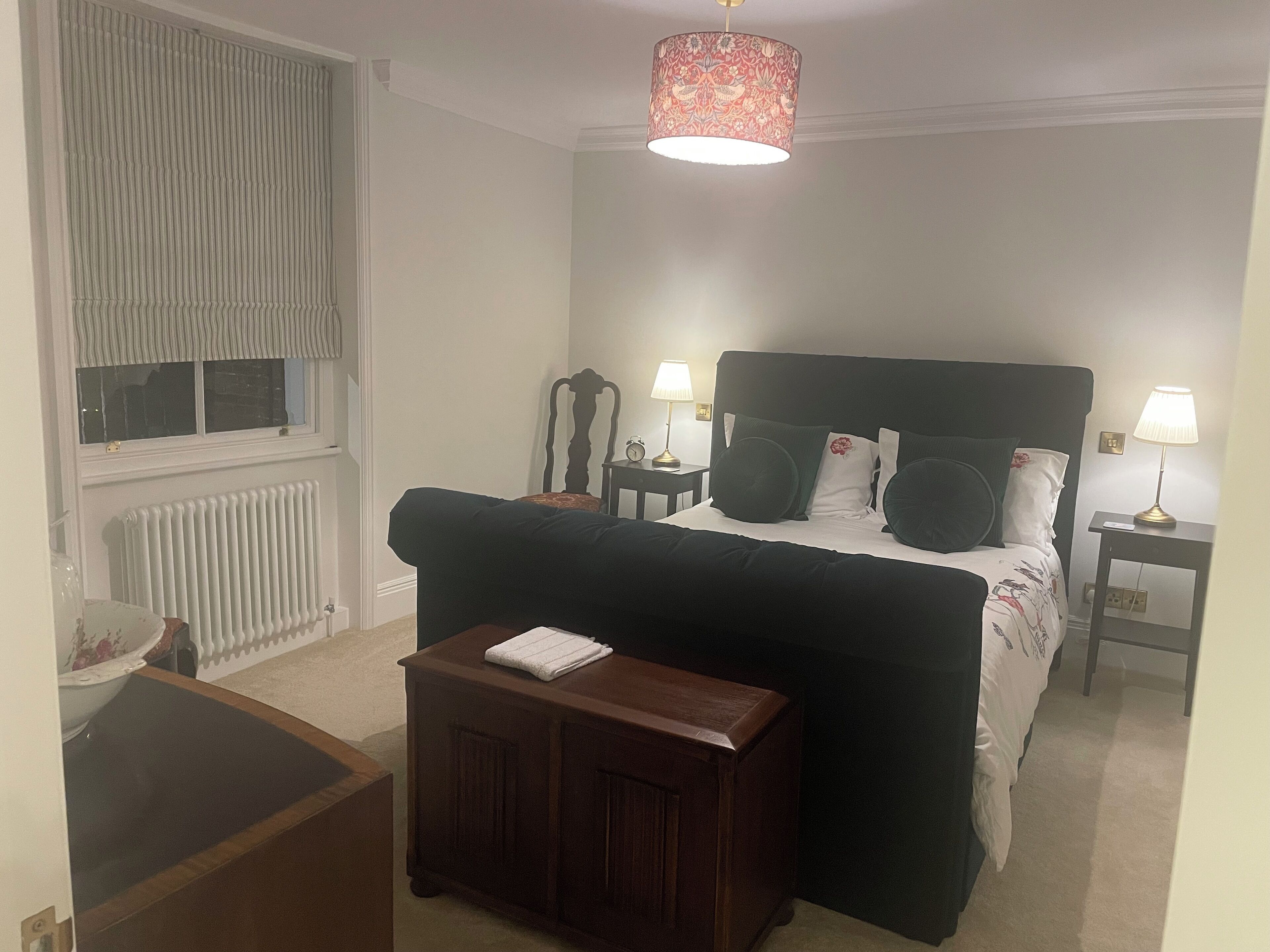 A bedroom with a bed, bedside tables, a nightstand, a wooden bench, a radiator, a window with curtains, and a hanging lamp.