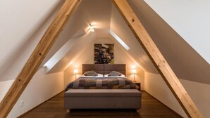 Un dormitorio con techo inclinado, vigas de madera, una cama con colcha estampada, dos lámparas de noche y un cuadro enmarcado en la pared.