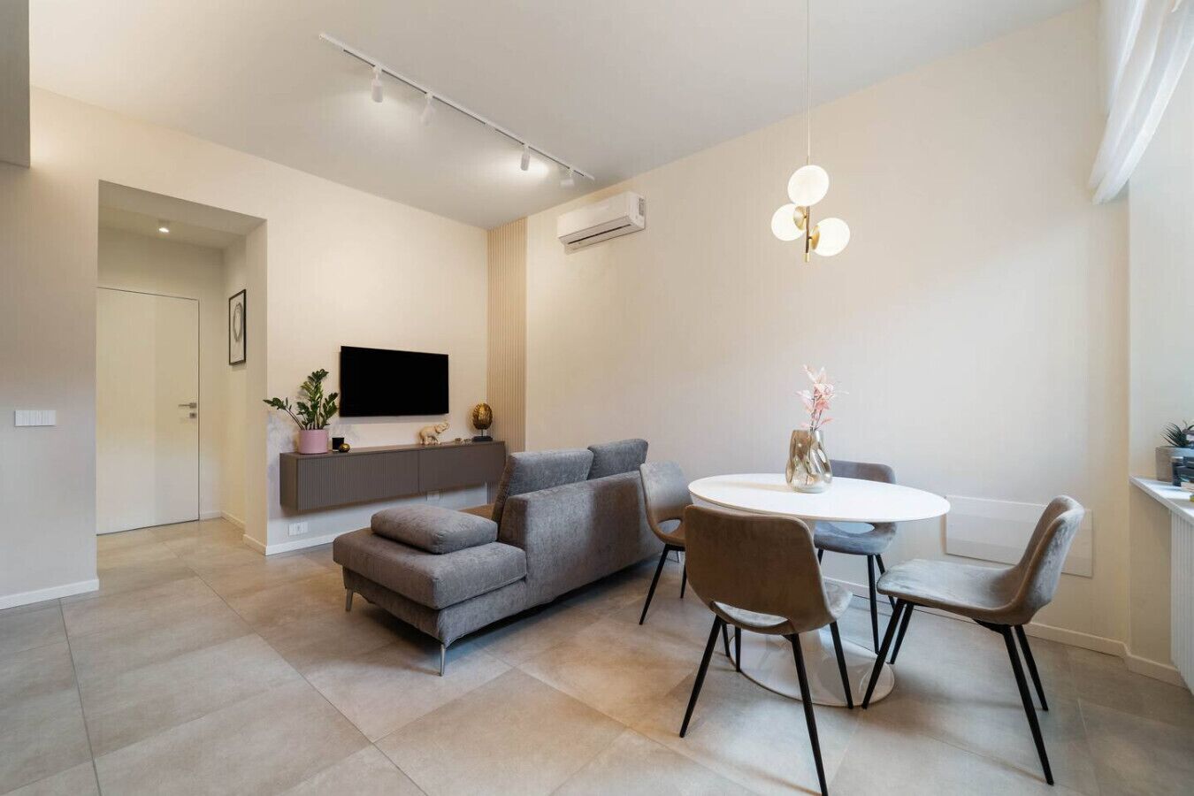 Ein modernes Wohnzimmer mit einem grauen Sofa, einem runden Esstisch mit vier Stühlen, einem flachbildfernseher an der Wand und einer Pendelleuchte über dem Tisch.