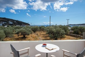 A terrace with a table, chairs, and a book. The terrace overlooks a landscape with olive trees and a distant view of the sea.