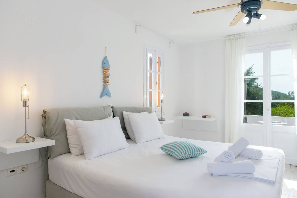 Una camera da letto con un letto, cuscini, un comodino con una lampada, una ventola a soffitto e una finestra con tende.