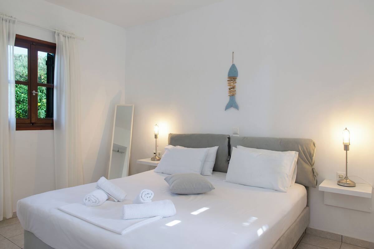 Un letto rifatto con lenzuola e asciugamani bianchi, un comodino con una lampada e una decorazione a forma di pesce appesa al muro.