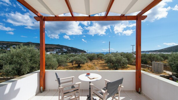 A covered outdoor seating area with a table and chairs, overlooking a landscape with olive trees and distant hills. - Blue Fish (Sifnos)