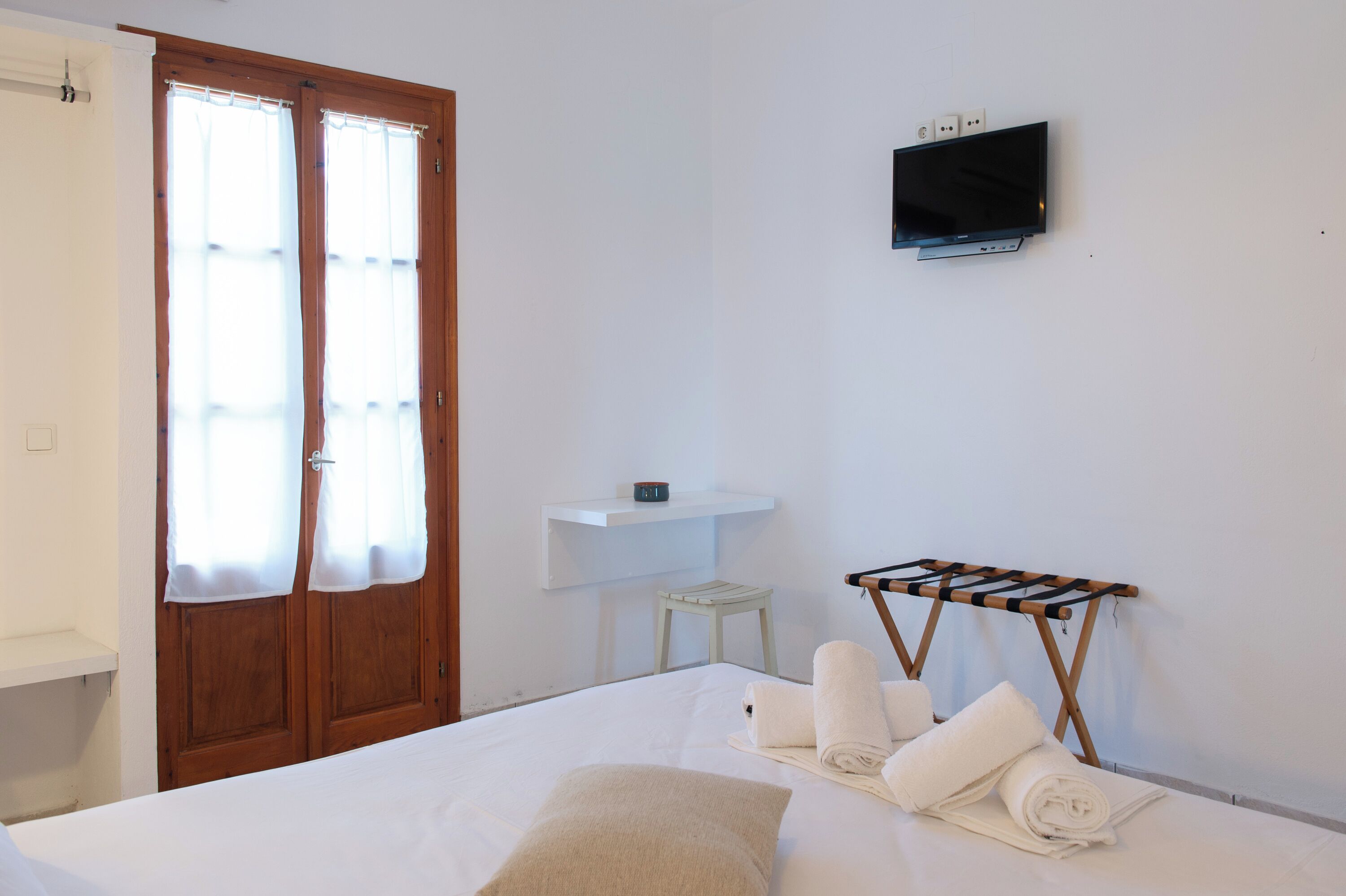 Une chambre d’hôtel avec un lit, un téléviseur fixé au mur, une petite étagère blanche et un étendoir en bois avec des serviettes.