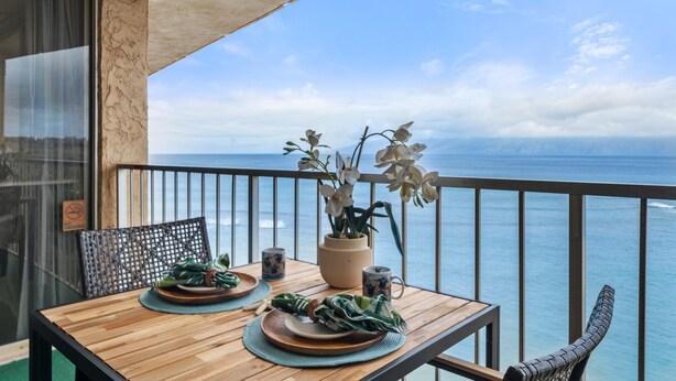 A wooden table with plates, a vase with flowers, and a chair on a balcony with a view of the ocean. - SUMMER SALE! Oceanfront 1BD Condo with Sunset & Whale Views - Royal Kahana 1206 (Lahaina)