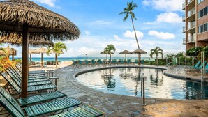 A pool area with lounge chairs and umbrellas, a palm tree, and a building in the background. - SUMMER SALE! Oceanfront 1BD Condo with Sunset & Whale Views - Royal Kahana 1206 (Lahaina)