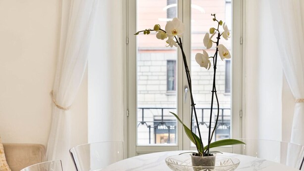Een eettafel met een glazen schaal met orchideeën, voor een raam met witte gordijnen.