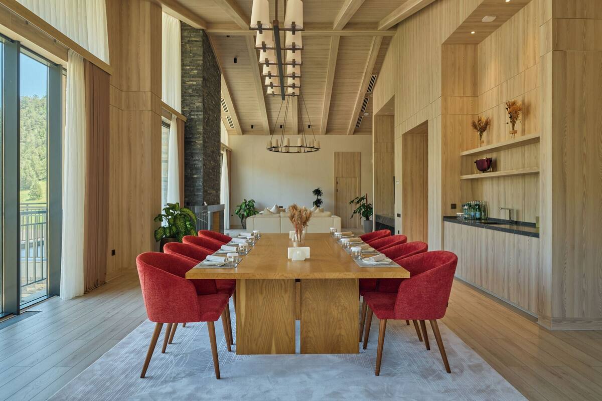 A spacious dining area with a long wooden table, red upholstered chairs, and a modern chandelier.