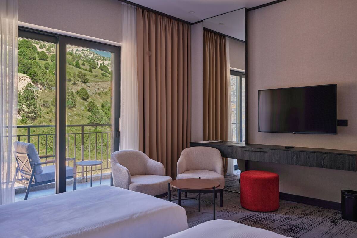 A hotel room with two beds, a TV, a desk, and a balcony with a chair and table.