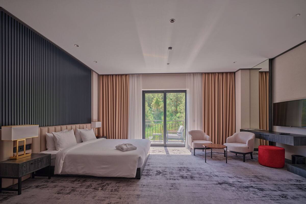 A modern hotel room with a large bed, a desk, a TV, and a balcony with curtains.