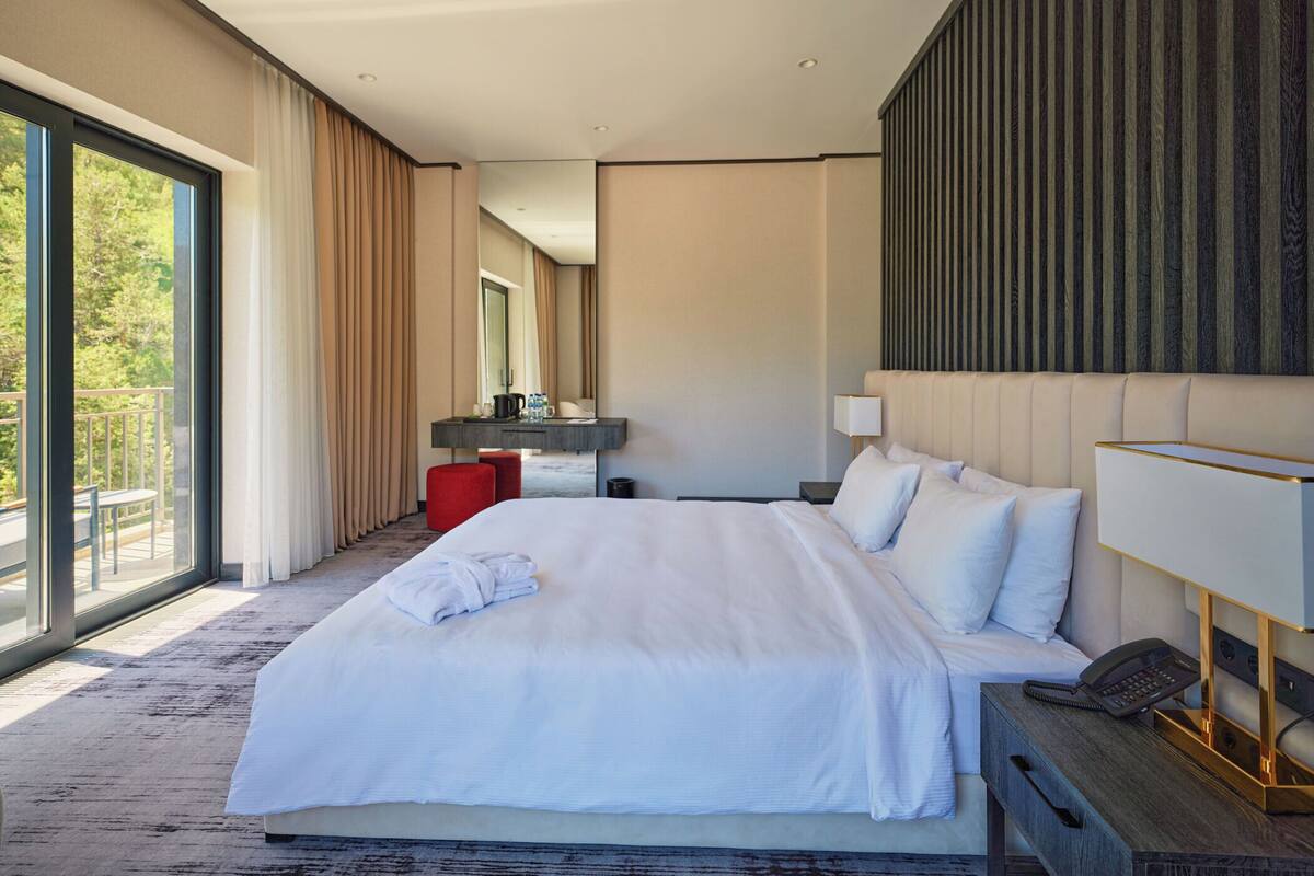 A modern hotel room with a large bed, a desk, a mirror, and a view of the outdoors.