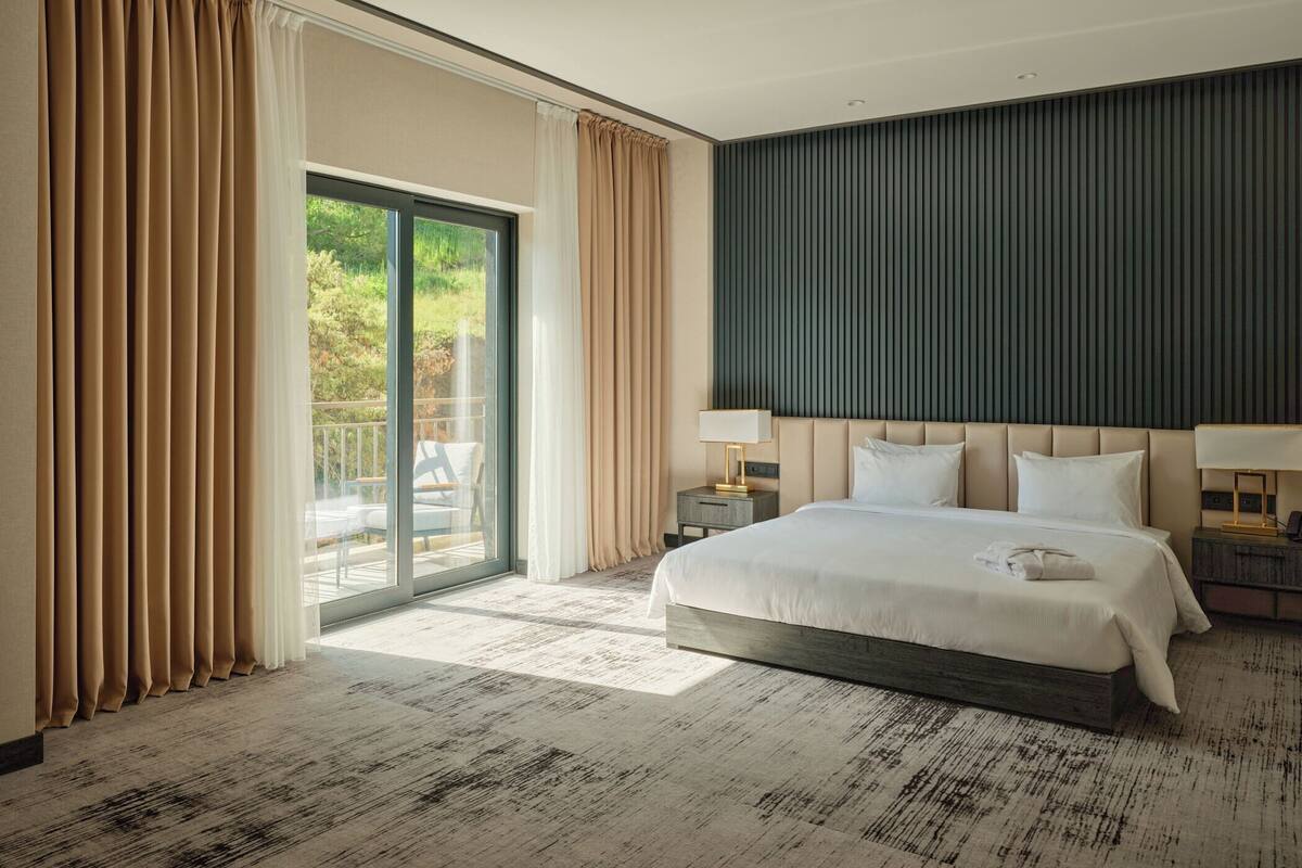 A modern hotel room with a large bed, a view of greenery outside, and a sliding glass door.