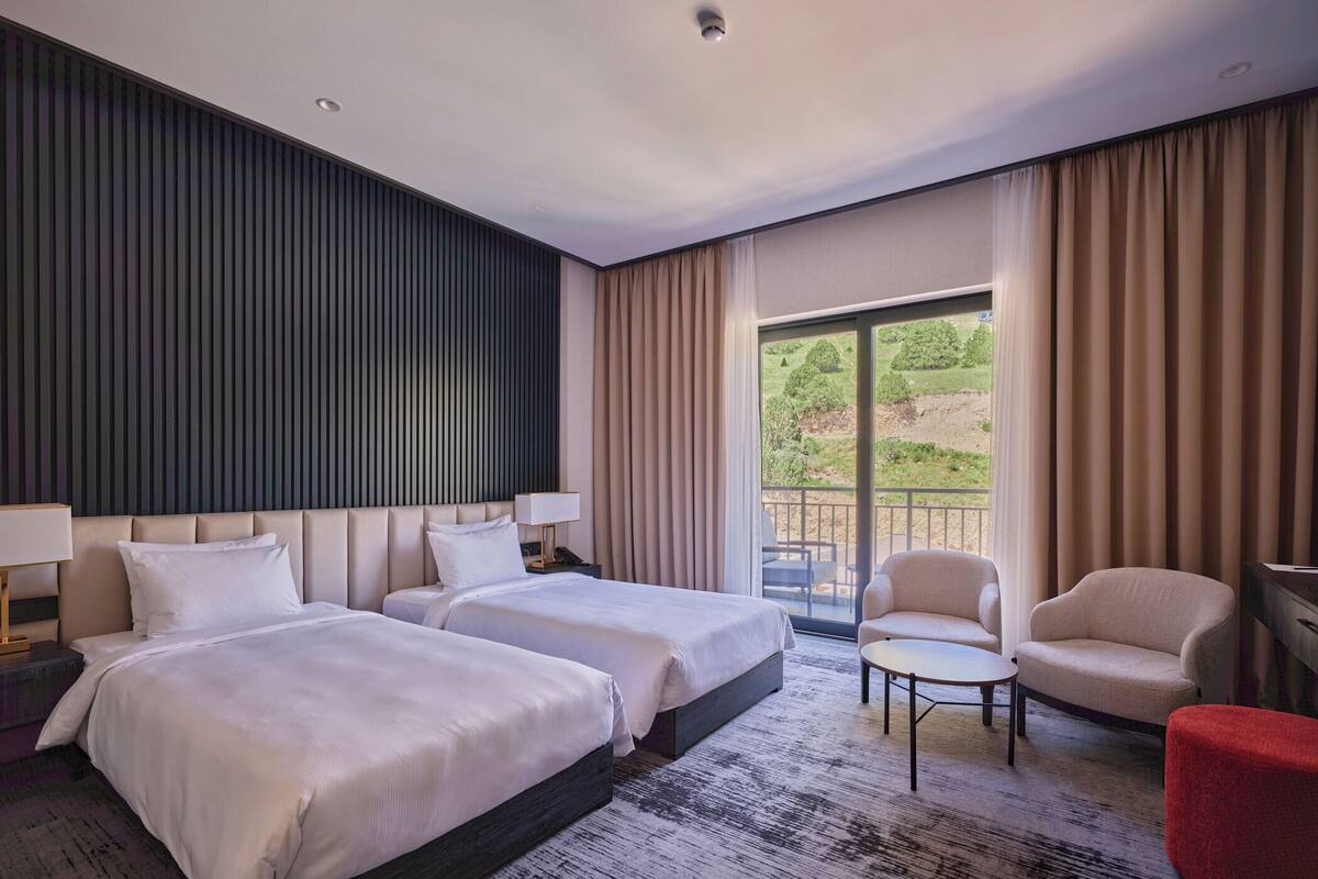 A hotel room with two beds, a small table, a chair, and a balcony with a view of greenery.