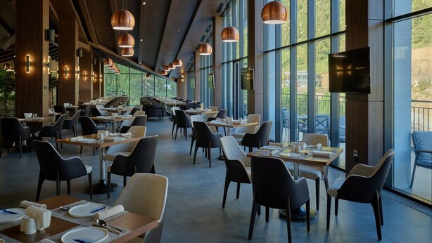 A spacious restaurant with high ceilings, large windows, and modern pendant lighting. Tables are neatly arranged with chairs, and a flat-screen TV is mounted on the wall. - Zamindor Resort (Zaamin)