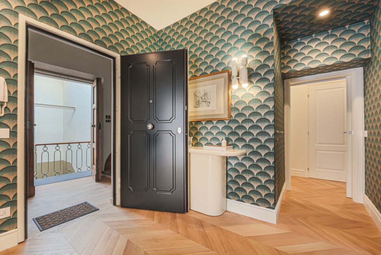 Un couloir avec un papier peint à motifs, une porte noire, un lavabo blanc et un tableau encadré accroché au mur.