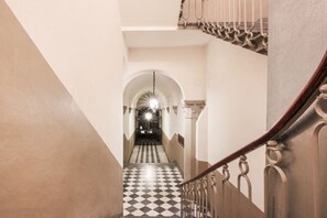 A hallway with a checkered floor, a staircase with a wooden handrail, and an arched doorway at the end. - Apartments Florence - The Four Suites (Firenze)