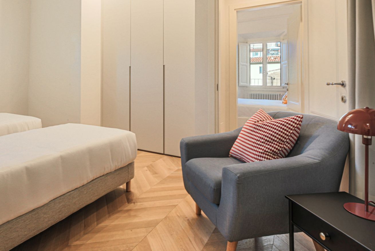 A bedroom with a bed, a grey armchair with a patterned pillow, a black side table, and a view of a balcony with a railing.