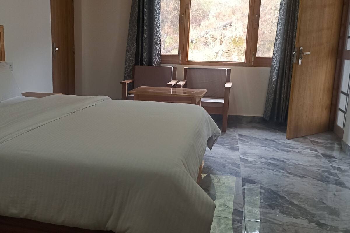 Ein Schlafzimmer mit einem Bett, einem Holzkopfteil, einem kleinen Tisch, zwei Stühlen und einem Fenster mit Vorhängen.
