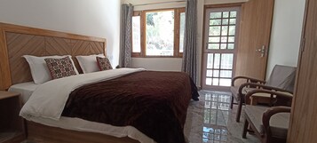 Un dormitorio con cabecera de madera, una cama grande con almohadas estampadas, un edredón rojo oscuro, una mesita de noche de madera, una silla y una puerta con ventanal.