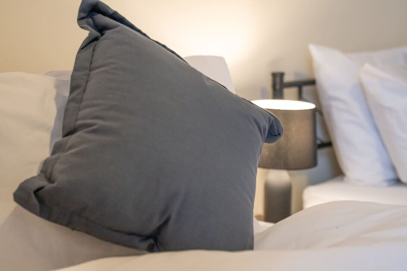 Una cama con un almohadón gris y una lámpara de noche.